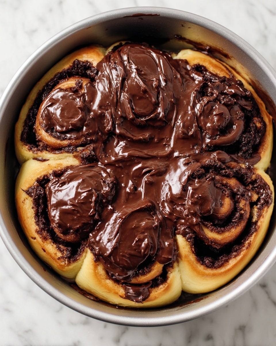 A round metal baking pan holds a batch of five cinnamon rolls tightly placed next to each other. Each roll has a light golden dough base with dark brown cinnamon filling visible in the spirals. A thick layer of shiny, smooth chocolate glaze covers the top of the rolls, pooling slightly in the creases and giving a rich contrast to the lighter dough underneath. The pan is set on a white marbled surface. The photo taken with an iphone --ar 4:5 --v 7