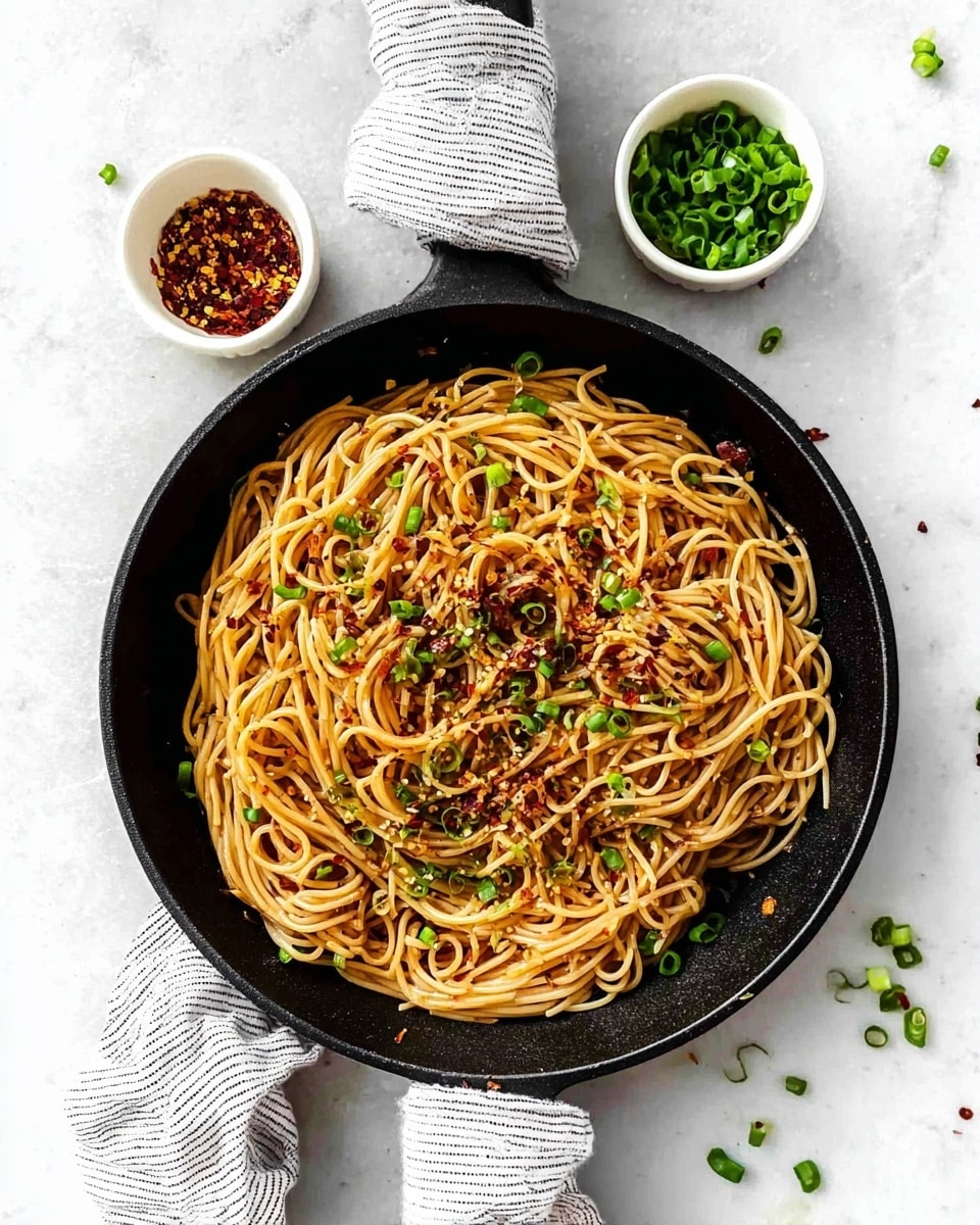 A black skillet filled with a single layer of light brown noodles, evenly mixed with small green chive pieces and red chili flakes on top. The skillet handle is wrapped with a white and gray striped cloth. Two small white bowls sit near the skillet on a white marbled textured background; one bowl has chopped green chives, and the other has red chili flakes. Some chive pieces are scattered around the skillet. Photo taken with an iphone --ar 4:5 --v 7