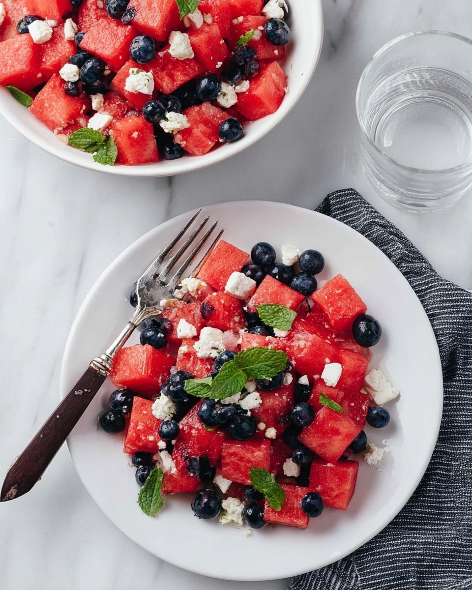 The image shows a white plate with a fresh fruit salad made of bright red watermelon cubes mixed with small round dark blueberries, scattered with small white cheese pieces and green mint leaves on top, placed next to a silver fork with a dark wooden handle on a black and white striped cloth. Above the plate, there is a white bowl filled with the same fruit salad, both placed on a white marbled surface. To the right of the bowl, a clear glass of water is visible. The colors are bright and fresh, with the red, blue, white, and green standing out against the clean white background and plate. photo taken with an iphone --ar 4:5 --v 7