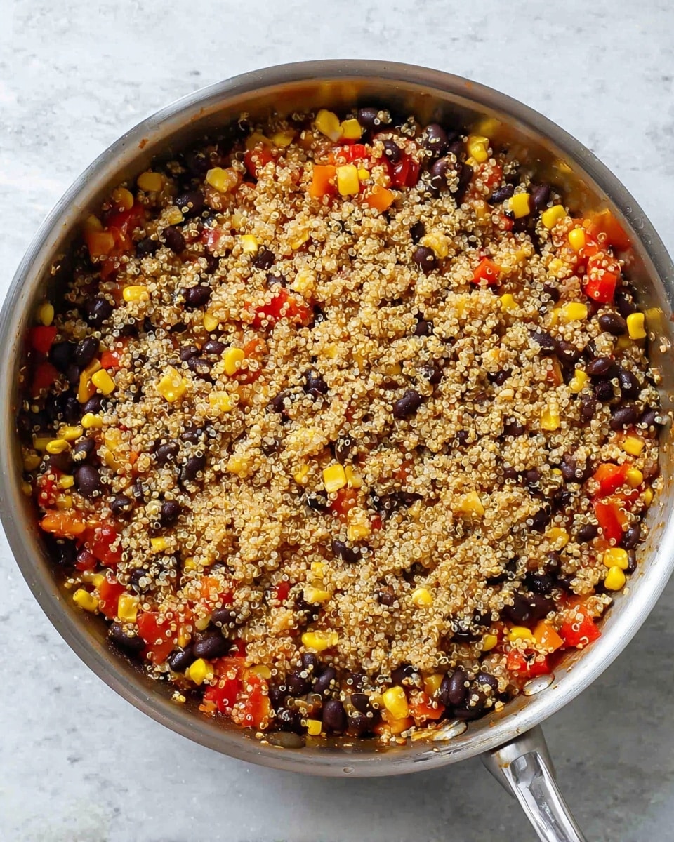 A silver frying pan filled with a cooked mixture that has two visible layers. The bottom layer consists of black beans, bright yellow corn kernels, and small pieces of red bell pepper all mixed together. The top layer is a thick cover of cooked quinoa, light beige in color with black quinoa seeds mixed in, giving a speckled texture. The pan handle shines brightly on the right side, and the pan sits on a white marbled surface. photo taken with an iphone --ar 4:5 --v 7