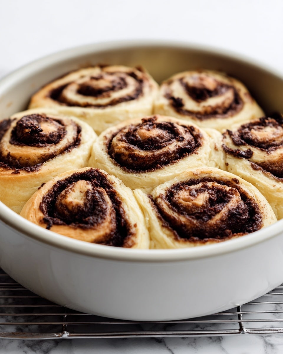 The image shows a round white baking pan filled with six swirled rolls made of light tan dough and dark brown filling. Each roll has visible spiral layers, with a thick dark brown layer rolled inside the soft light tan dough. The rolls are closely packed, touching each other and filling the pan. The pan is placed on a metal cooling rack over a white marbled surface, and the background is a clean white. photo taken with an iphone --ar 4:5 --v 7