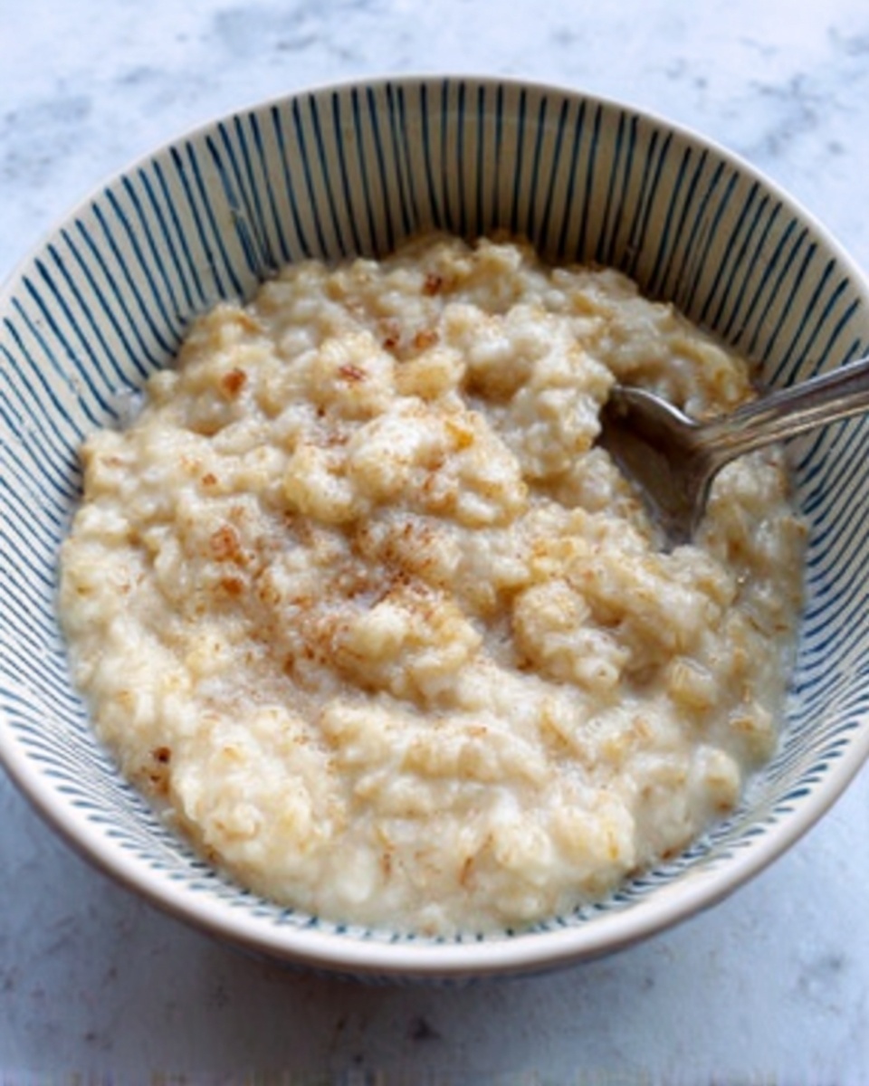 A white bowl with thin blue vertical lines inside holds thick, creamy oatmeal with a soft, lumpy texture, light beige in color with small brown bits mixed in. A metal spoon rests inside the bowl on the right side. The bowl is placed on a white marbled surface. photo taken with an iphone --ar 4:5 --v 7