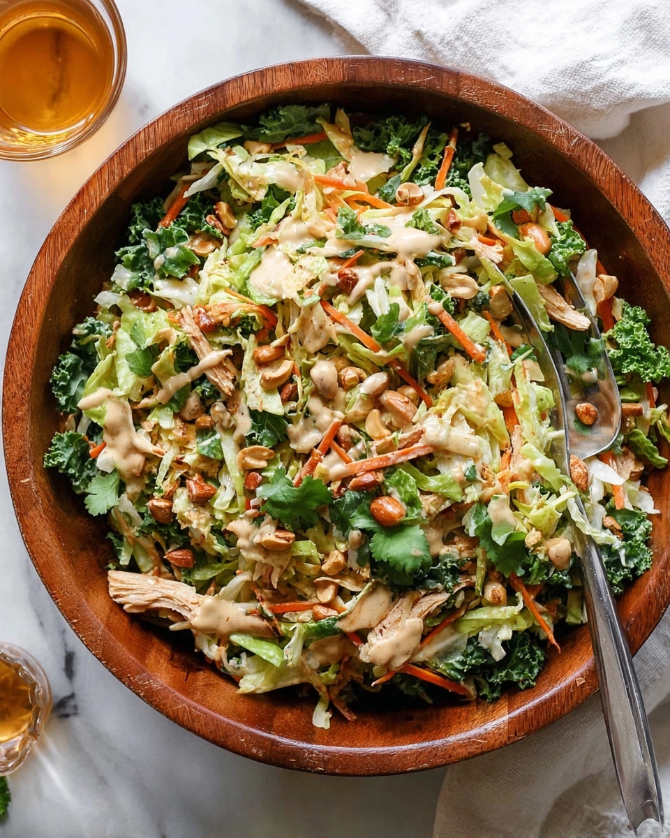 A wooden bowl filled with a colorful salad showing many layers starting from shredded green and light yellow cabbage, mixed with thin orange carrot sticks and dark green kale leaves. Scattered on top are golden brown nuts and shredded light beige chicken pieces, with fresh green cilantro leaves adding extra color. The salad is lightly covered with a creamy beige dressing that is drizzled unevenly across the top. A silver fork is visible inside the bowl, and the whole setup is on a white marbled surface with a white cloth and a glass cup of light amber liquid partly visible to the side. Photo taken with an iphone --ar 4:5 --v 7