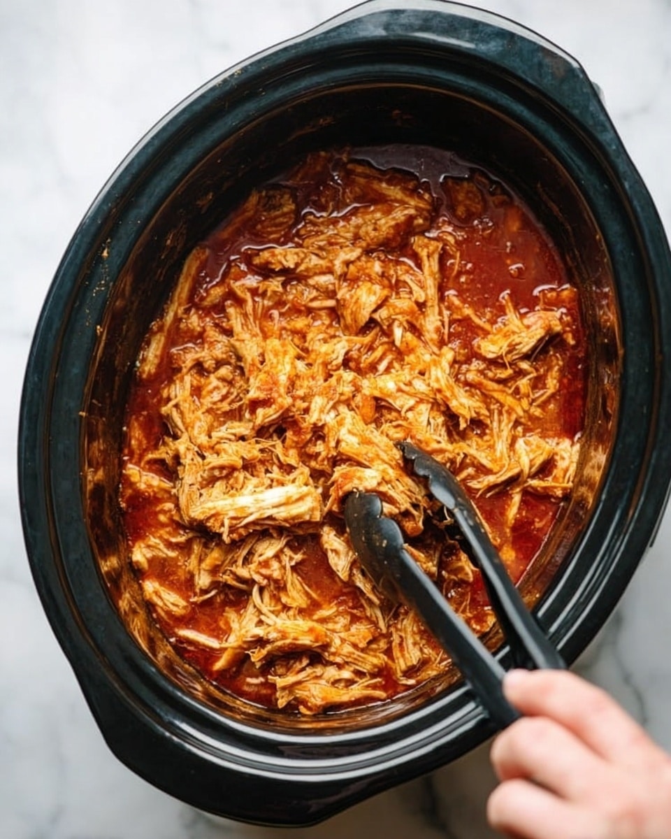 The image shows a slow cooker filled with shredded cooked chicken in a thick red sauce. The chicken is soft and stringy, sitting in the bottom of the black cooker. The sauce looks rich and slightly oily, covering the chicken evenly. A pair of black tongs held by a woman's hand is lifting some of the shredded chicken from the cooker. The slow cooker is on a white marbled surface. photo taken with an iphone --ar 4:5 --v 7