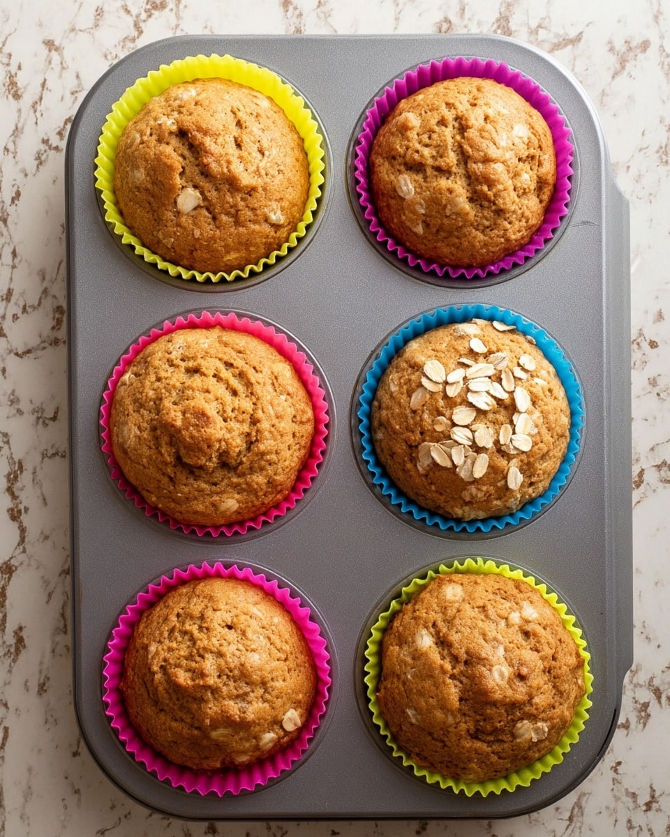 Six light brown muffins with soft, slightly cracked tops sit evenly placed inside colorful silicone liners—two yellow, two pink, one blue, and one purple—each liner holding one muffin. Two muffins have oat flakes lightly sprinkled on their tops, adding texture and a hint of off-white color. The muffins rest in a gray metal muffin tray with six round cups, all placed on a white marbled surface with subtle veins of gray and beige. The view is from above, showing the round shapes and gentle golden tones of the muffins clearly. photo taken with an iphone --ar 4:5 --v 7