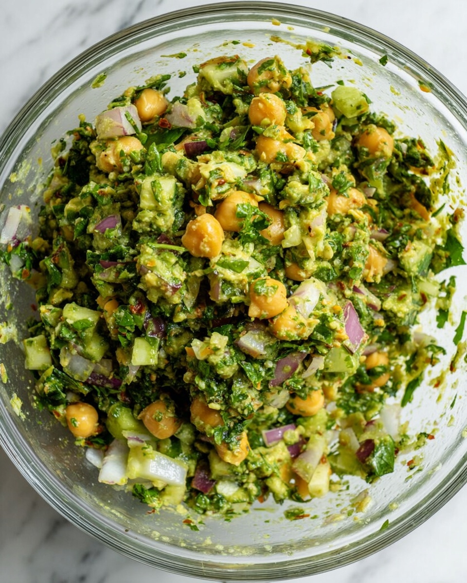 A clear glass bowl filled with a mixture of finely chopped green vegetables and herbs, combined with small yellow chickpeas and bits of red onion, all coated evenly in a green sauce or dressing; the ingredients are roughly chopped, showing varying textures and colors throughout the salad; it rests on a white marbled surface. photo taken with an iphone --ar 4:5 --v 7