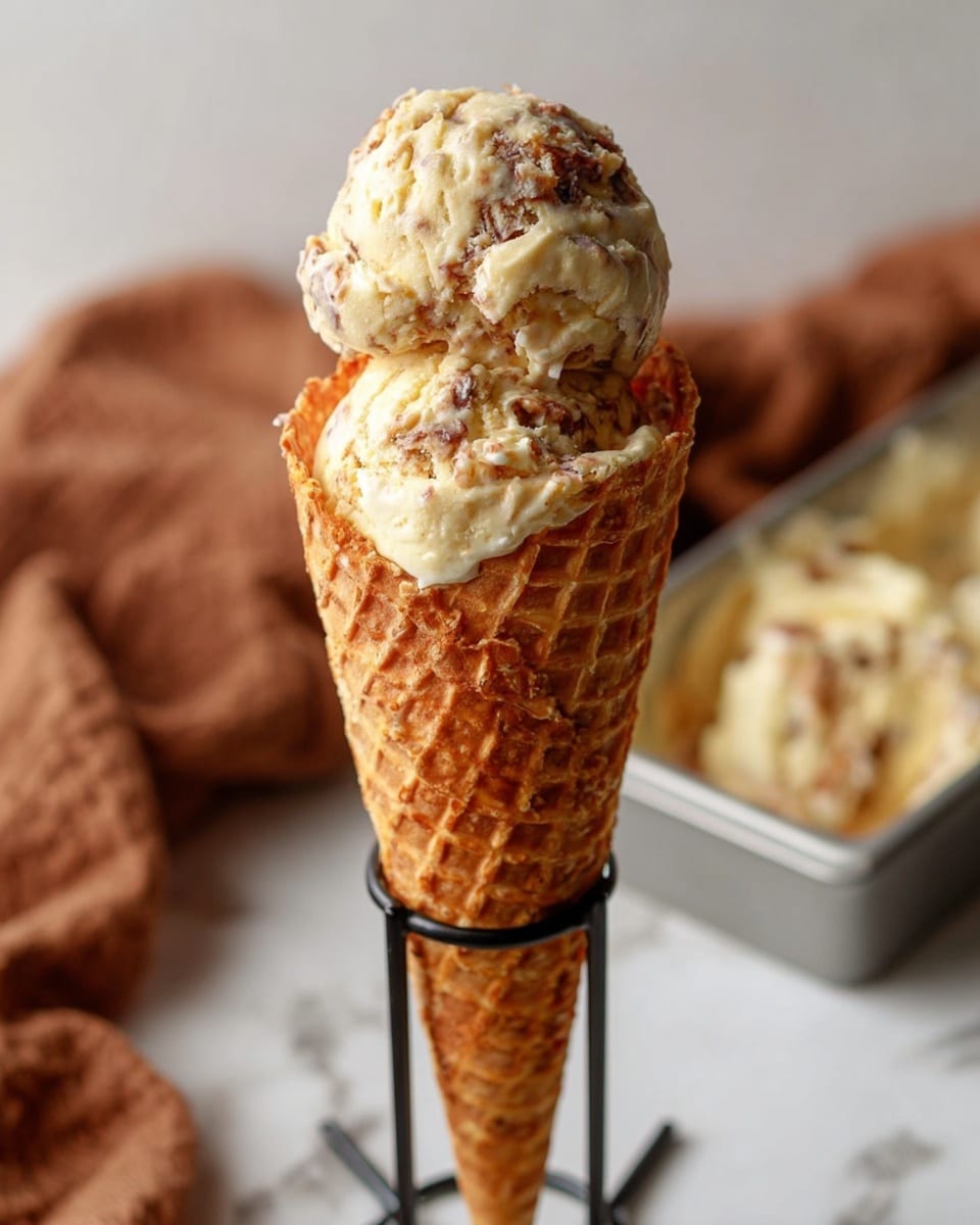 A tall ice cream cone with two large scoops of creamy ice cream that has swirls of brown mixed in, sitting on top of a crisp, golden waffle cone with a textured grid pattern. The cone is held upright by a black metal holder on a white marbled surface, with a soft brown cloth blurred in the background and a silver tray partially visible on the side holding more ice cream. The overall look is warm and inviting, showcasing the textured ice cream and the crunchy cone. Photo taken with an iphone --ar 4:5 --v 7