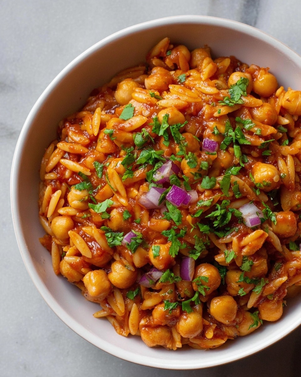 A white bowl filled with a mix of small round chickpeas and orzo pasta coated in a red-orange sauce, garnished with chopped green cilantro and small pieces of purple onion scattered on top and mixed through. The chickpeas are plump and round, the orzo is smooth and slightly curved, all sitting on a plain white marbled background. photo taken with an iphone --ar 4:5 --v 7