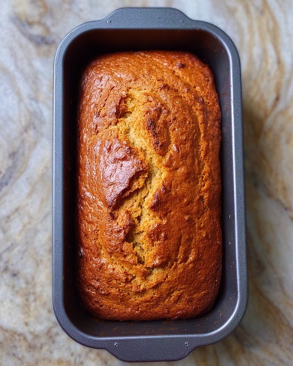 A freshly baked loaf of bread with a golden-brown crust sits still in a dark gray rectangular baking pan. The top of the bread shows natural cracks and a lightly textured surface, indicating a soft inside. The pan is placed on a white marbled surface that adds a clean and bright contrast to the warm loaf and dark pan. photo taken with an iphone --ar 4:5 --v 7