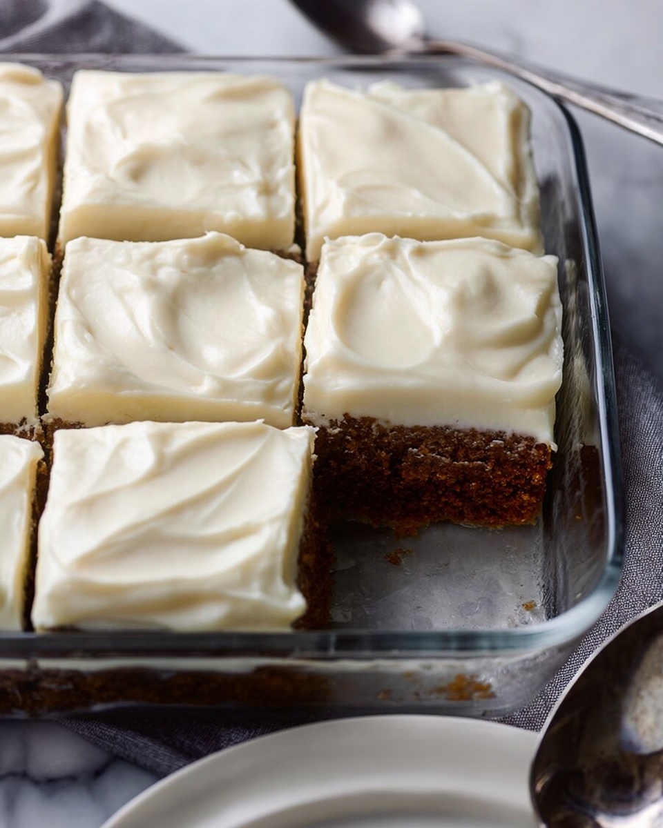 A glass dish contains six square pieces of a two-layer dessert. The bottom layer is brown and crumbly, resembling a cake or bar base. The top layer is a thick, smooth white frosting spread evenly with soft swirled textures. One piece is slightly lifted, showing the thickness of the layers. The dish rests on a white marbled surface, next to part of a white plate and a silver serving spoon on a light cloth. Photo taken with an iphone --ar 4:5 --v 7
