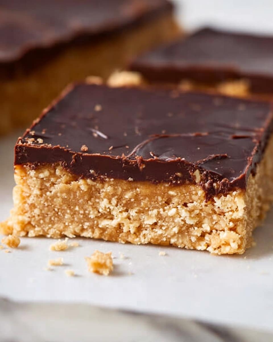 A close-up of a two-layer square dessert bar on a white marbled surface. The bottom layer is thick, crumbly, and light tan with a coarse texture, resembling a cookie or oat base. The top layer is a smooth, glossy dark chocolate sheet that covers the entire surface evenly, with slight cracks and subtle markings. There are small crumbs scattered around the bar on the white marbled surface. Photo taken with an iphone --ar 4:5 --v 7