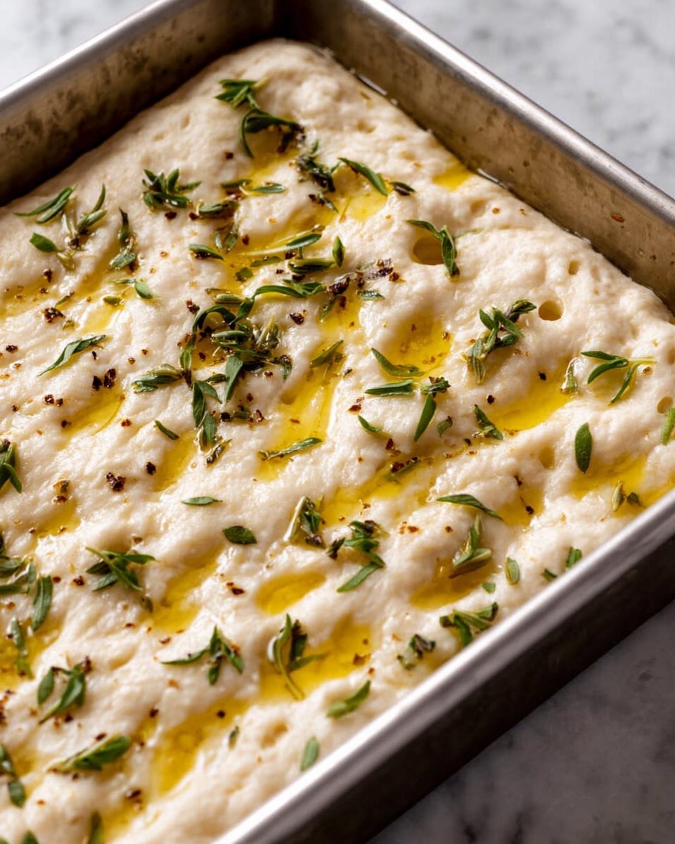 A close-up view of a white dough in a rectangular metal pan, showing a thick, bubbly texture with small deep indentations on the surface. The dough is lightly drizzled with golden olive oil that pools in the dimples, and small green herb leaves are scattered evenly across the top, adding bursts of color and texture. The metal pan rests on a white marbled surface. photo taken with an iphone --ar 4:5 --v 7