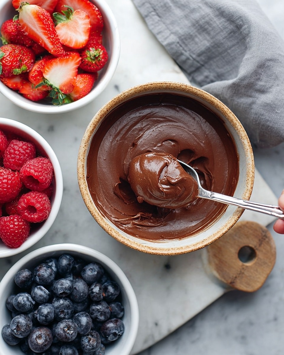 A clear bowl filled with smooth, dark brown chocolate pudding sits in the center, with a silver spoon resting inside and coated in the pudding. Around the bowl are three small white bowls: one holding bright red strawberries, some whole and some sliced, another with deep blue blueberries, and the third filled with vivid red raspberries. All bowls and the chopping board beneath have a clean white marbled texture background, creating a fresh look. A woman's hand is adjusting the spoon inside the bowl. photo taken with an iphone --ar 4:5 --v 7