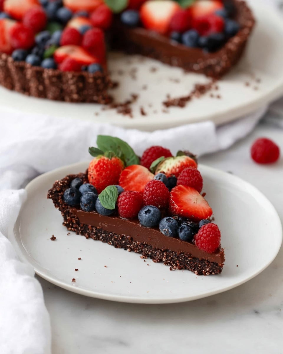 A slice of tart sits on a white plate on a white marbled surface, showing three layers: the bottom layer is a thick, textured dark brown crust that looks crunchy, the middle layer is a smooth, rich chocolate filling in dark brown, and the top layer is decorated with fresh red strawberries, red raspberries, and dark blue blueberries, some with green leaves attached, giving a colorful and fresh look. In the background, the whole tart is visible with a missing slice, and some crumbs are scattered on the plate and surface. A white cloth is softly placed near the plate. photo taken with an iphone --ar 4:5 --v 7