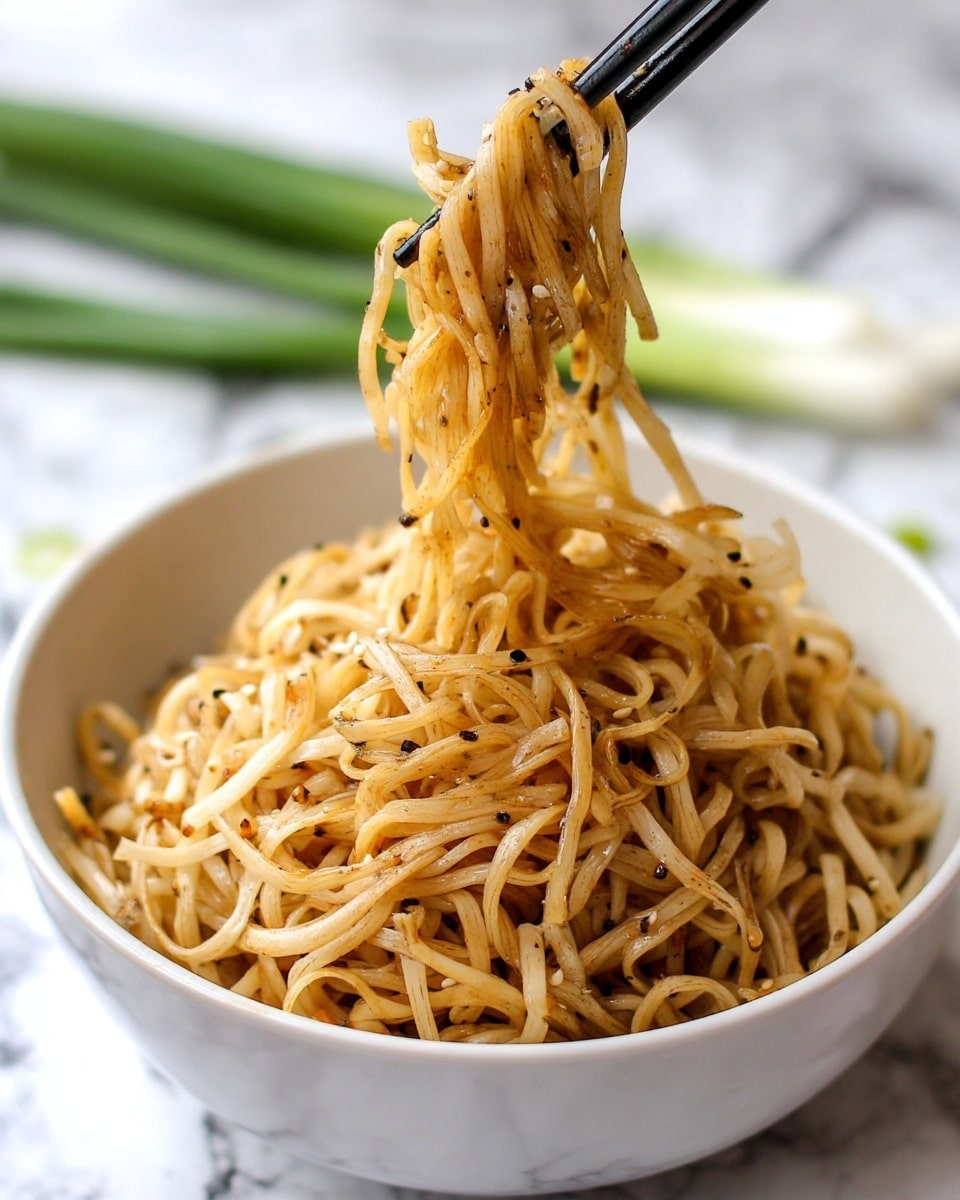 A white bowl filled with a large heap of thin, light brown noodles mixed with small black pepper specks and tiny sesame seeds. The noodles look soft and tangled, with a slightly oily texture that makes them shine. A pair of black chopsticks is holding a small bundle of noodles above the bowl, lifting them as if ready to eat. The surface underneath is a white marbled texture with some green spring onions visible in the background. photo taken with an iphone --ar 4:5 --v 7