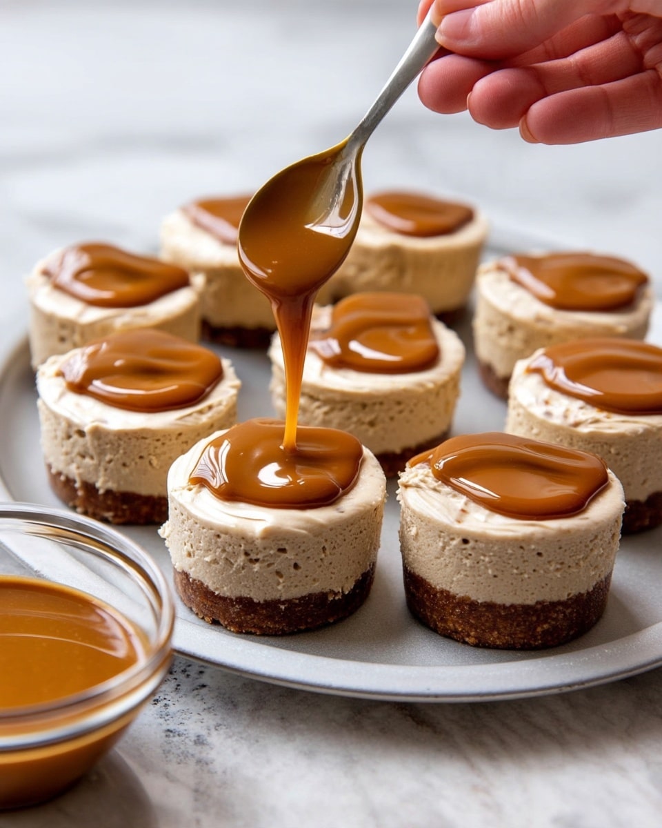 Mini cheesecakes are arranged on a round silver tray atop a white marbled surface. Each cheesecake has two layers: a firm brown base and a thick, creamy light tan top layer with a smooth but slightly textured surface. A woman's hand holds a spoon above one cheesecake, pouring a shiny, smooth caramel-colored sauce in the center of the top layer, creating soft curves as it spreads. In the bottom left corner, a clear bowl filled with the same caramel sauce is partially visible. Photo taken with an iphone --ar 4:5 --v 7